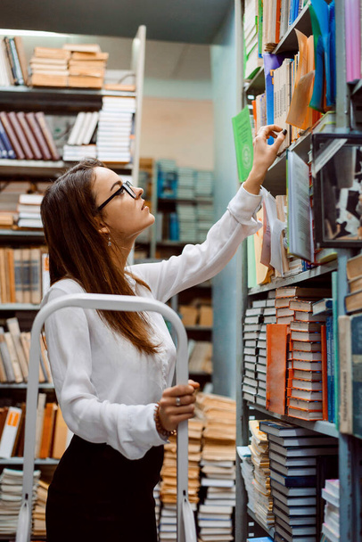 Όμορφη κοπέλα-βιβλιοθηκάριος κανονίζει βιβλία στα ράφια. Έννοια της βιβλιοθήκης - Φωτογραφία, εικόνα