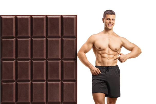 Shirtless man leaning on a big chocolate and showing abdominal muscles isolated on white background - Foto, Imagen