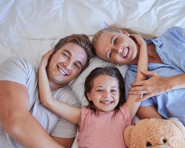 Adorable little girl pulling her parents close while lying in between her mother and father, from above. Faces of loving parents bonding with their daughter lying on a bed and spending free time toge. - Valokuva, kuva