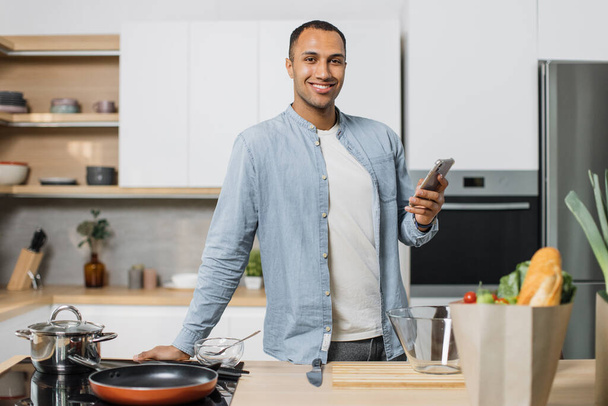 Happy young indian man using smartphone standing in kitchen in the morning. Happy hindu guy reading news and enjoying leisure time at home, empty copy space. - Foto, Bild