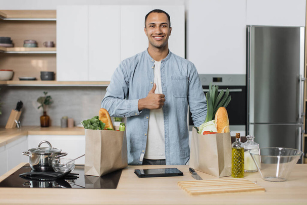Young positive attractive indian man preparing salad, healthy food, from fresh organic vegetables, showing thumb up standing on the background of new modern light kitchen at home. - Photo, image
