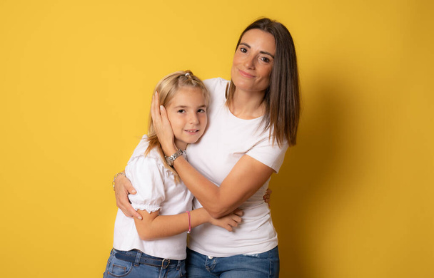 Beautiful mother and daughter smiling happy hugging over isolated yellow background. - Photo, Image