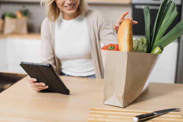 Cropped view of happy woman cooking healthy food looking on paper bag with vegetables while reading recipe on digital tablet in modern kitchen at home on sunny day. Preparing vegetable salad. - Foto, Bild