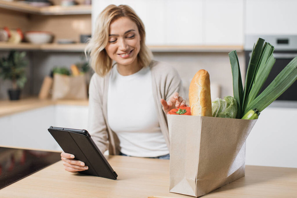 Focus on hand holding tablet and paper bag with vegetables. Happy woman cooking healthy food looking on paper bag with vegetables while reading recipe on digital tablet in modern kitchen at home. - Foto, Bild