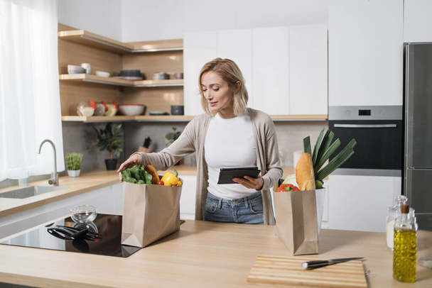 Positive cheerful young blond woman, searching menu recipe of healthy vegetarian vegetable salad in digital tablet in modern bright kitchen at home. Smiling female, food and technology concept. - Foto, Bild