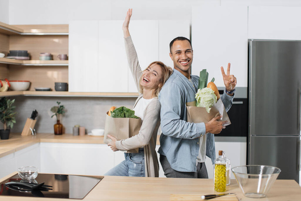 Positive joyful young indian man and blond woman standing together in kitchen, smiling, standing back to back and unpacking grocery paper bag. Concept of family, domestic lifestyles and healthy eating - 写真・画像