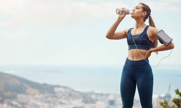 One fit young mixed race woman taking a rest break to drink water from bottle while exercising outdoors. Female athlete wearing earphones and armband quenching thirst and cooling down after running a. - Photo, Image
