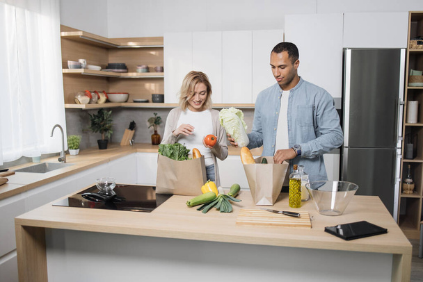 Positive young people in love unpacking shopping back with fresh organic products. Caucasian family standing together on kitchen and enjoying domestic chores. - 写真・画像