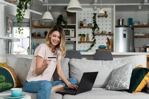 Smiling happy woman paying online, using laptop, holding plastic credit card, sitting on couch at home, positive young female shopping, making secure internet payment, browsing banking service - Foto, Bild