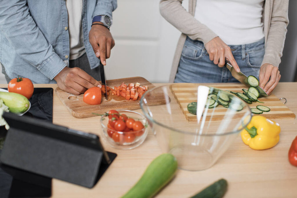 Close up of hands cutting fresh vegetables tomato and cucumber with knife, couple preparing salad. Unrecognizable man and woman cooking dinner together and having fun in a new modern apartment. - Photo, image
