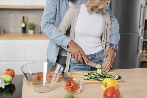 Genç ve çekici çiftin taze sebzelerden salata hazırlarken çekilmiş görüntüsü. Yakışıklı sporcu ve sarışın kadın birlikte yemek pişiriyor ve modern bir dairede eğleniyorlar.. - Fotoğraf, Görsel