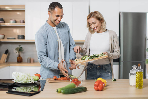 Happy couple standing in modern kitchen cooking salad breakfast or dinner together. Smiling young husband and wife vegetarians chopping vegetables and dropping them into a glass bowl preparing lunch - Photo, image