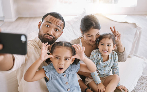 Portrait of a mixed race family taking a selfie and making funny faces while sitting on the sofa at home. Hispanic man using a smartphone to take a photo with his wife and two little cute daughters i. - Fotoğraf, Görsel