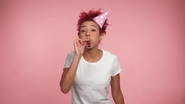 Happy African American redhead curly woman in birthday cap blowing whistle celebrating holiday. Joyful positive smiling female wearing white t-shirt on isolated pink background with copy space - Footage, Video