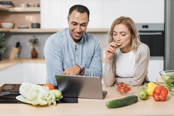 Handsome indian man and caucasian blond woman using laptop searching recipes, ordering shopping online, watching cooking class during preparing delicious vegan salad from fresh vegetables. - Foto, imagen
