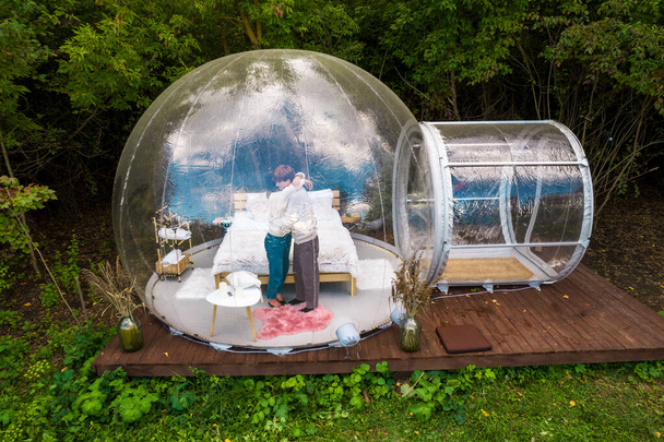 Tente à bulles transparente au glamping, forêt luxuriante autour et à l'intérieur. Un couple qui se serre dans ses bras - Photo, image