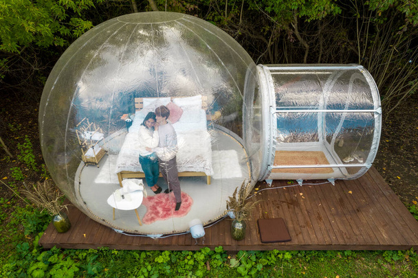 Transparentní bublina stan na glamping, svěží les kolem a interiér. Pár objímající se navzájem - Fotografie, Obrázek