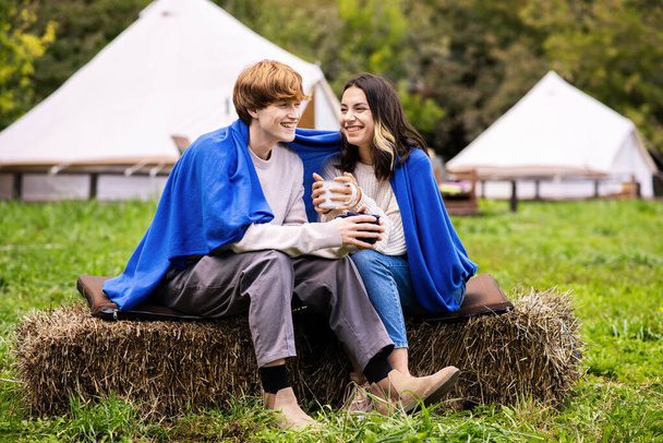 A couple sitting under a blanket on a stump and holding cups at glamping. Tents on the background, lush forest around - Фото, изображение