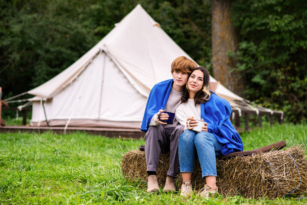 A couple sitting under a blanket on a stump, holding cups and hugging each other at glamping. Tents on the background, lush forest around - Фото, зображення