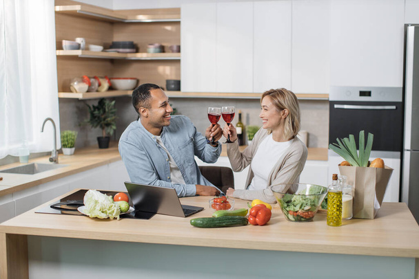 Joyful family cooking dinner at modern light home. Couple in love, caucasian blond woman and handsome smiling arab man talking and making salad, drinking red wine and enjoy perfect day. - Photo, image