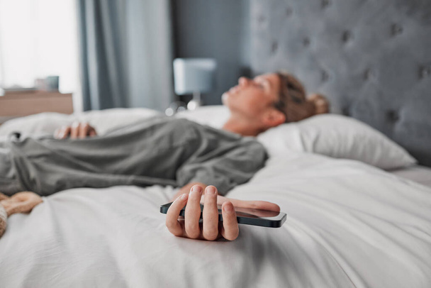 Phone, bed and woman sleeping in a bedroom, exhausted and suffering with mental health problem. Tired, lazy and procrastination by depressed lady ignoring alarm, experience fatigue from insomnia. - Foto, Imagem