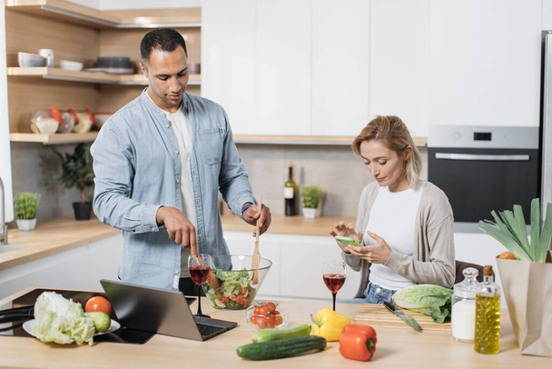 Portrait of loving multinational couple cooking salad together on a kitchen. Cheerful blond woman using mobile phone to find recipe of vegan salad while her husband mixing vegetables in a glass bowl - Zdjęcie, obraz