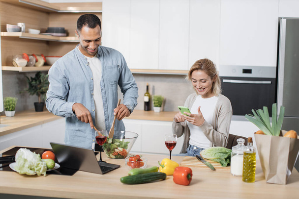 Portrait of loving multinational couple cooking salad together on a kitchen. Cheerful blond woman using mobile phone to find recipe of vegan salad while her husband mixing vegetables in a glass bowl - Photo, Image