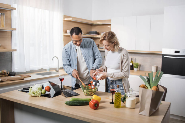 Young attractive couple in love preparing salad from fresh vegetables. Handsome sporty man mixing slices of ingredients and blond charming woman pouring olive oil into a glass bowl. - Valokuva, kuva