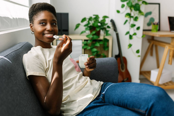 Young african woman relaxing on sofa eating organic yogurt. Diet nutrition with slim woman tasting healthy breakfast or bio snack product at home - Фото, изображение