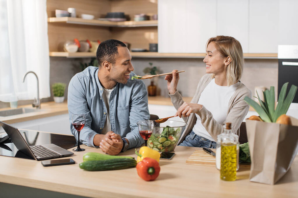 Χαρούμενο παντρεμένο πολυεθνικό ζευγάρι που χρησιμοποιεί φορητό υπολογιστή ενώ μαγειρεύει υγιεινό φαγητό στην κουζίνα, ξανθές νεαρές συζύγους που διασκεδάζουν ενώ ταΐζουν τον άντρα της με ξύλινη κουτάλα με σαλάτα από φρέσκα λαχανικά - Φωτογραφία, εικόνα