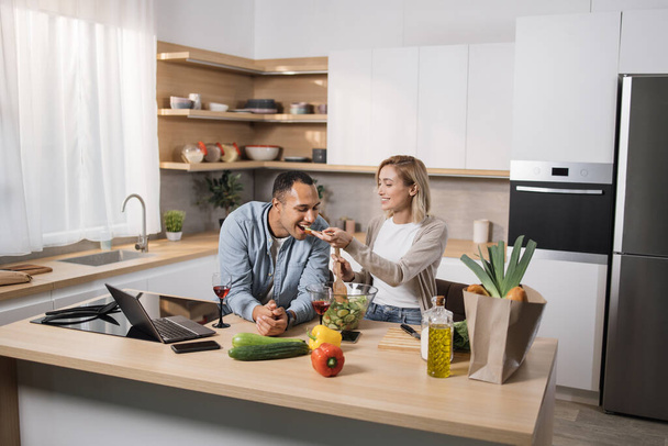 Cheerful married multinational couple using laptop while cooking healthy food in kitchen, blond young wife spouses having fun while feeding her husband wooden spoon with salad from fresh vegetables - Foto, Bild