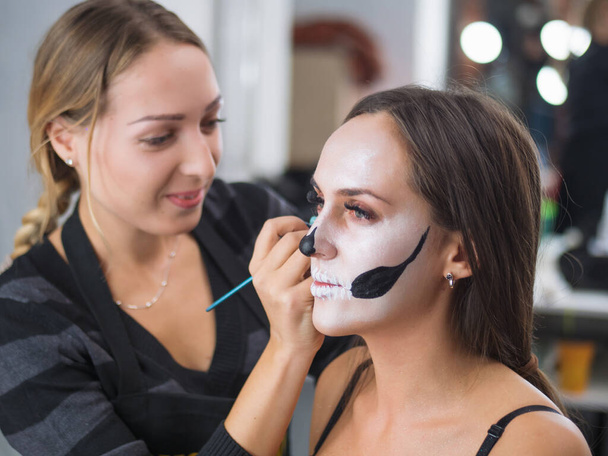 Make-up artist makes girl halloween skull glamour make up. Face art. Woman applies on professional greasepaint on face of lady. . High quality photo - Photo, Image