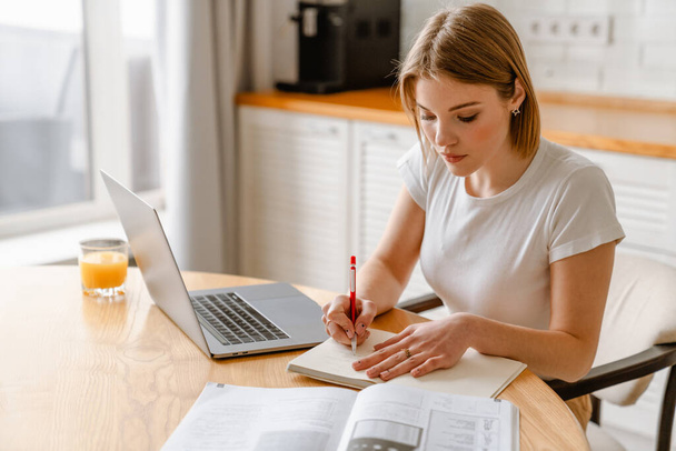 Pensive jonge blonde witte vrouw zitten met laptop computer aan de keukentafel het maken van notities - Foto, afbeelding