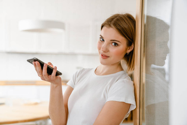 Smiling young woman leaning on a wall in the kitchen at home, holding mobile phone - Photo, Image
