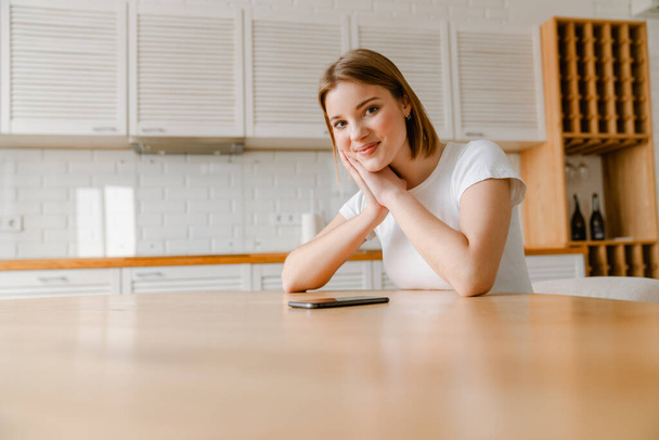Усміхнена молода жінка сидить за кухонним столом вдома з мобільним телефоном
 - Фото, зображення