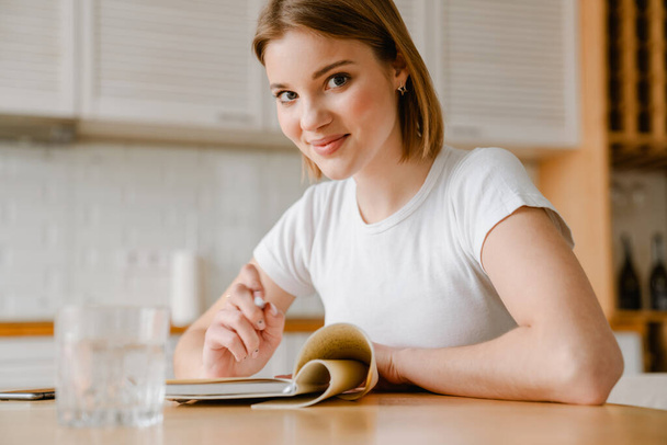 Smiling young woman writing in her diary while sitting at the kitchen table at home - Photo, image