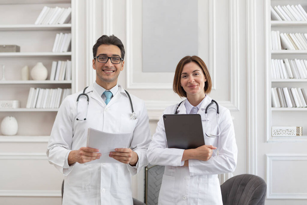 Foto de retrato de dos jóvenes doctores sonrientes en bata médica blanca en el consultorio de la clínica. Reunión de colegas, discusión y concepto de atención médica - Foto, Imagen