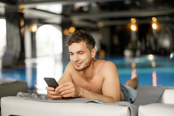 Man rests by the swimming pool indoors, talking on phone while lying relaxed on lounge bed indoors. Man spends holidays at spa resort with an indoor pool - Foto, Bild