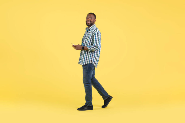 Chico negro usando Smartphone sonriendo a la cámara Websurf y mensajes de texto caminando posando en el estudio sobre fondo amarillo. Anuncio de aplicación móvil. Disparo de longitud completa - Foto, imagen