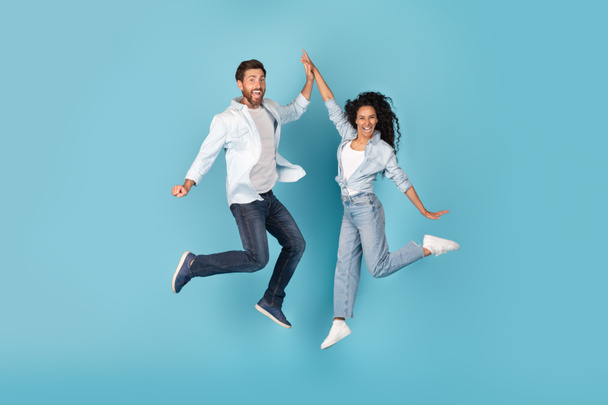 Excited cheerful millennial caucasian and arabic couple jumping in air, give high five, on free space, isolated on blue background, studio. Fun together, success and victory emotions, great ad, offer - Photo, Image