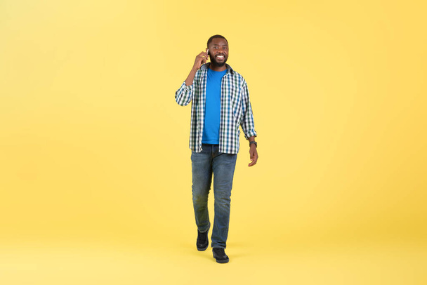 スタジオで黄色の背景にカメラに笑みを浮かべて歩くポーズ携帯電話で話す陽気な黒人男性.モバイル通信とガジェットの概念。フルレングスショット - 写真・画像