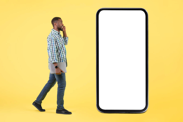 Czarny człowiek mówiący przez telefon komórkowy Walking Blisko Big Smartphone z pustym ekranem pozowanie trzymając laptopa komputera na żółtym tle. Reklama Oferty Mobilnej. Zdjęcia studyjne, makieta - Zdjęcie, obraz