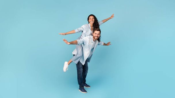 Cheerful millennial caucasian man holding middle eastern wife on his back, doing plane, have fun, isolated on blue background, studio. Ad and offer, love and relationship, couple enjoy freedom, love - Photo, Image