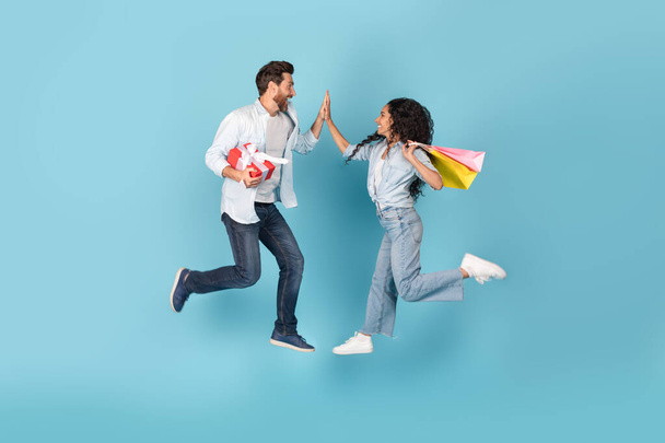 Excited glad millennial international couple with gift and bags jump in air, give high five on copy space, isolated on blue background, studio. Good ad and offer, fun together, success and win, sale - Photo, Image