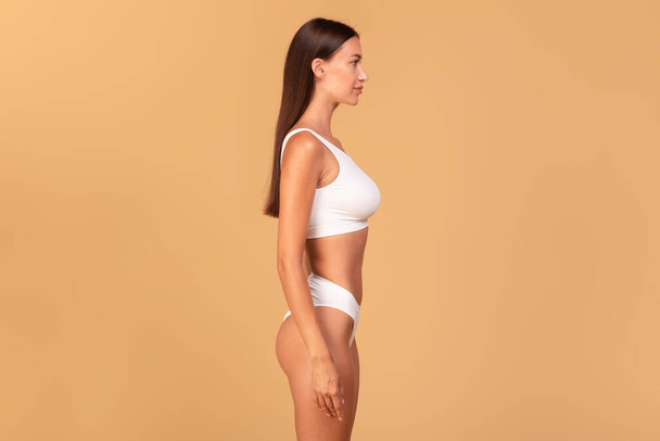 Side view of slim lady in white underwear showing beautiful body with smooth skin, young woman with sporty fit figure posing on beige studio background, free copy space - Photo, Image