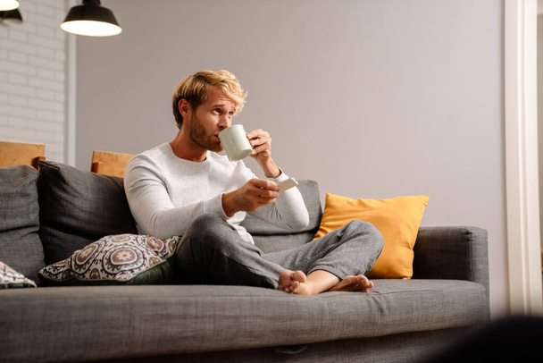 Blonde man drinking tea and watching tv while sitting on sofa at home - Fotoğraf, Görsel