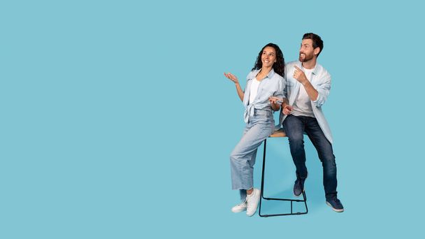 驚くべき幸せな千年の白人の夫とアラブの妻が一緒に椅子に座って、青い背景、スタジオに隔離された空の空間に指を表示します。選択、巨大な広告と提供し、人々の感情 - 写真・画像