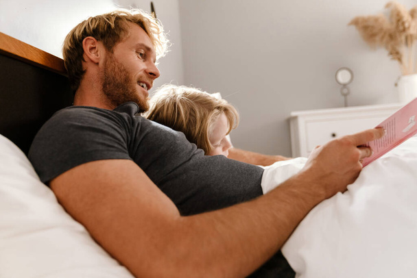 Pai branco e filho lendo livro enquanto descansam juntos na cama em casa - Foto, Imagem