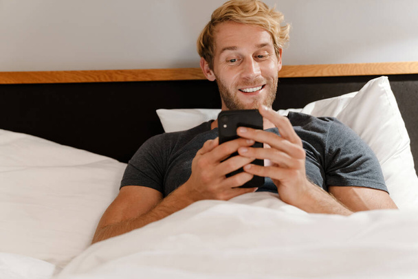 Blonde bearded man using mobile phone while lying in bed at home - Foto, Bild