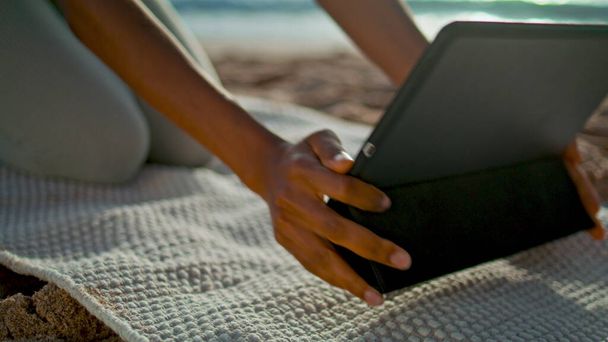 Unknown girl using tablet on beach close up. Young african american woman putting tab computer on yoga mat preparing to online training outdoors. Slim lady have modern gadget for workout on nature. - Foto, afbeelding